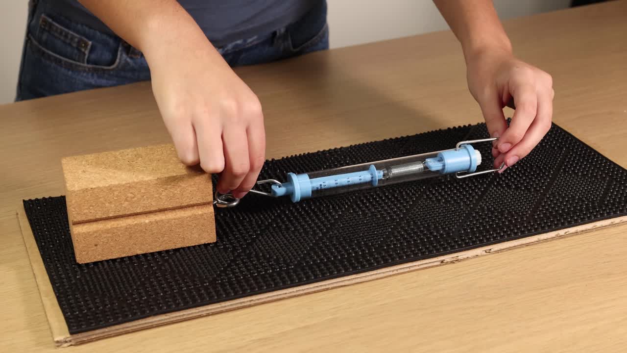 A wooden block is dragged across a surface using a spring scale, illustrating friction and force measurement