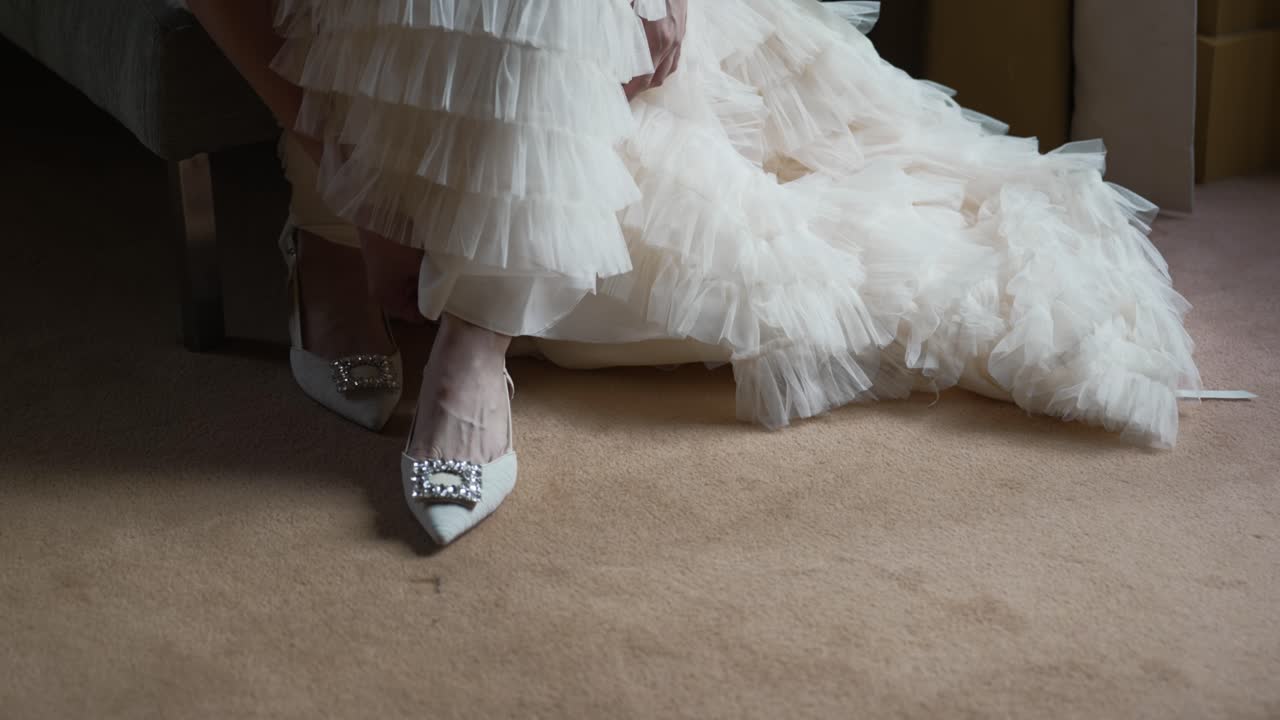 Bride in layered white gown adjusts jeweled heels in soft intimate moment before ceremony