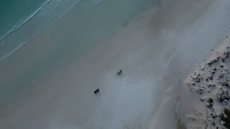 Two-vehicles-travelling-across-the-sand-at-the-seashore,-aerial-tracking-shot,-spotlight-mode