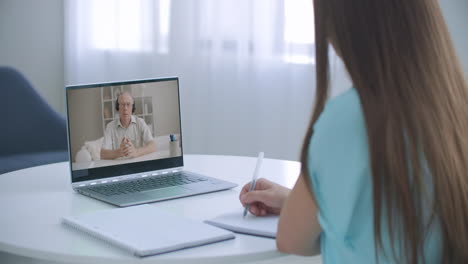 Girl-school-college-student-distance-learning-waving-hand-studying-with-online-teacher-on-laptop-screen.-Elearning-zoom-video-call-videoconference-class-with-tutor.-Over-shoulder-view.