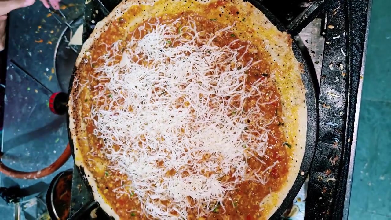 Lähikuva katukeittiön myyjästä raastamassa tuoretta juustoa masala dosan päälle kuumalla tawa-pannulla, valmistaen rapeaa eteläintialaista ruokalajia vilkkaalla intialaisella ruokakojulla.