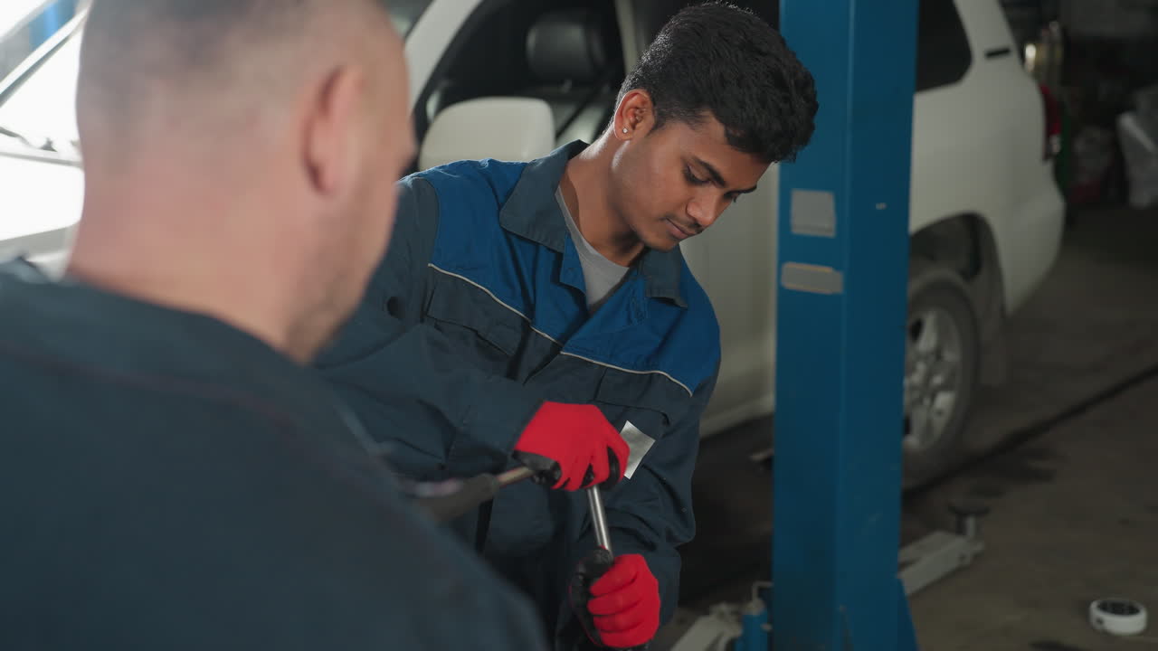 mecánico en uniforme azul y guantes rojos apretando pernos en el motor del coche mientras el colega observa desde atrás, el fondo presenta a alguien pasando en negro cerca de un ascensor de vehículo en un taller automotriz