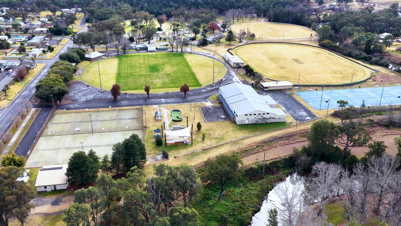 Aerial footage captures a smooth drone flyover above sports fields, tennis courts, and surrounding suburban streets in Coonabarabran, New South Wales, under daylight