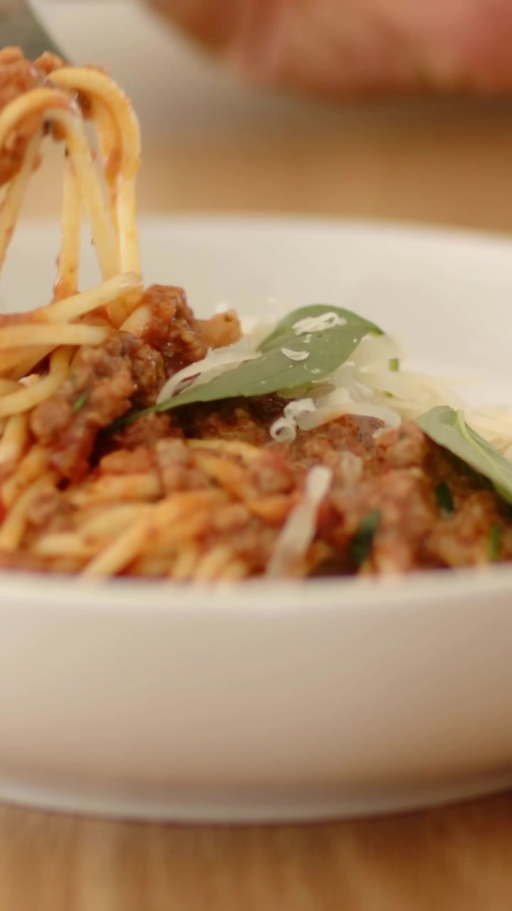 Video verticale: Mano che inserisce una forchetta che gira in una ciotola su un tavolo di legno, sollevando spaghetti verso la bocca.