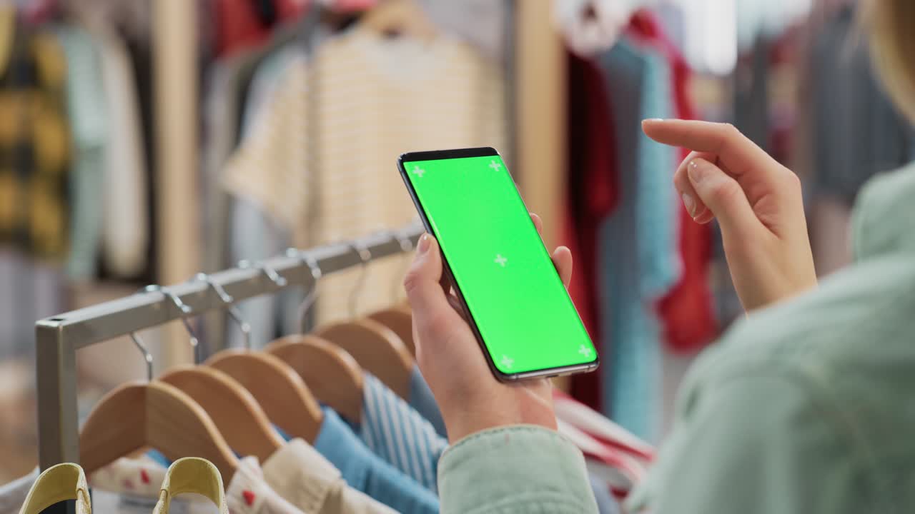 tienda de ropa: mujer usando teléfono inteligente con pantalla verde de clave de croma. colgante de ropa con elegantes artículos de marca para venta al por menor en el fondo. imágenes cercanas del dispositivo móvil.