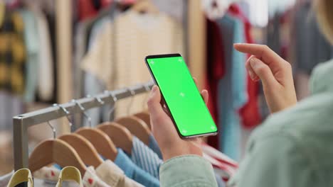 clothing store: female using smartphone with chroma key green screen display. clothes hanger with stylish branded items for retail sale in the background. close up footage of mobile device.