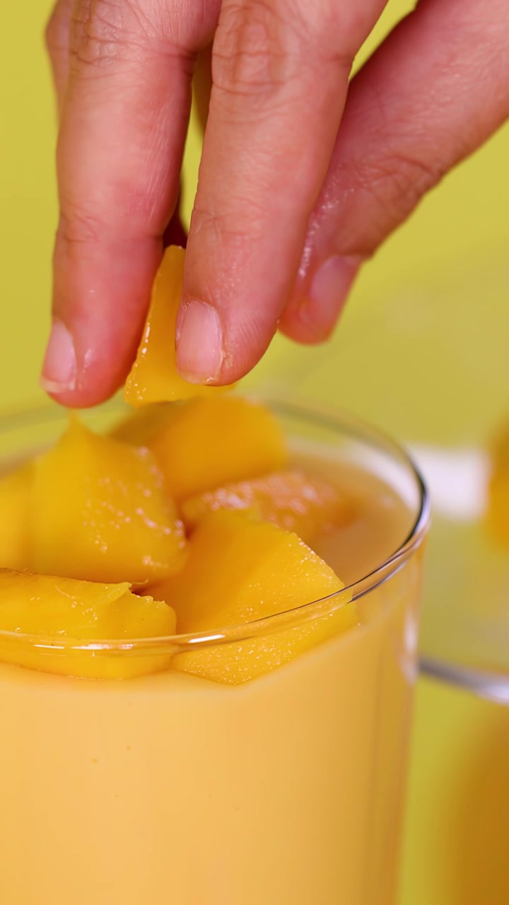 Primo piano di una mano che aggiunge cubetti di mango a un frullato cremoso sotto una brillante illuminazione da studio gialla.