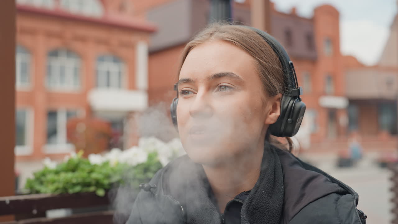 young woman with headphones outdoor cafe, thoughtful expressions, vaping device held to lips, relaxed posture on bench, brick storefront background, soft bokeh, warm autumn light, candid portrait