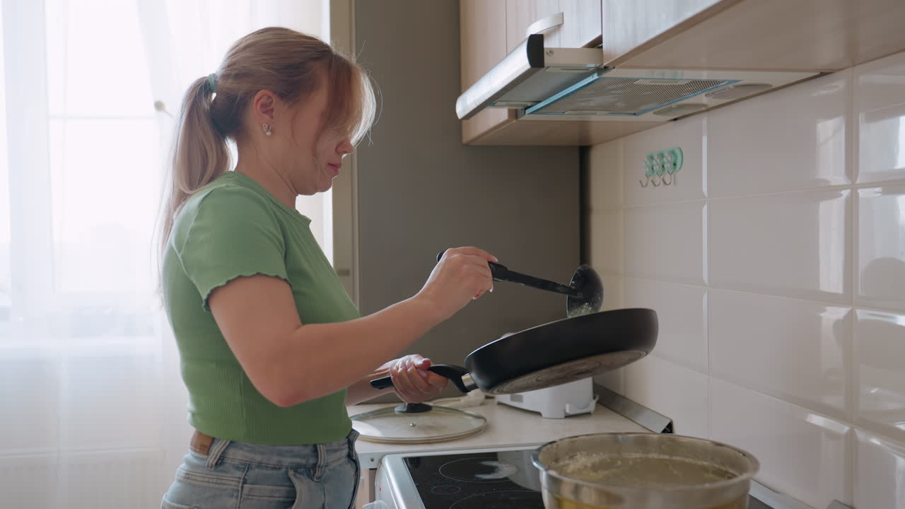 Woman pours pancake batter into frying pan on stove in bright kitchen, preparing homemade breakfast with care and focus, capturing cooking process and healthy lifestyle