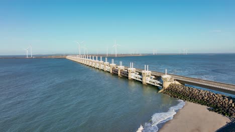 Toma-Aérea-En-Cámara-Lenta-De-Una-Barrera-De-Marejada-Ciclónica-Abierta-Del-Este-De-Scheldt-Y-Turbinas-Eólicas-En-Zelanda,-Los-Países-Bajos-En-Un-Hermoso-Día-Soleado-Con-Un-Cielo-Azul