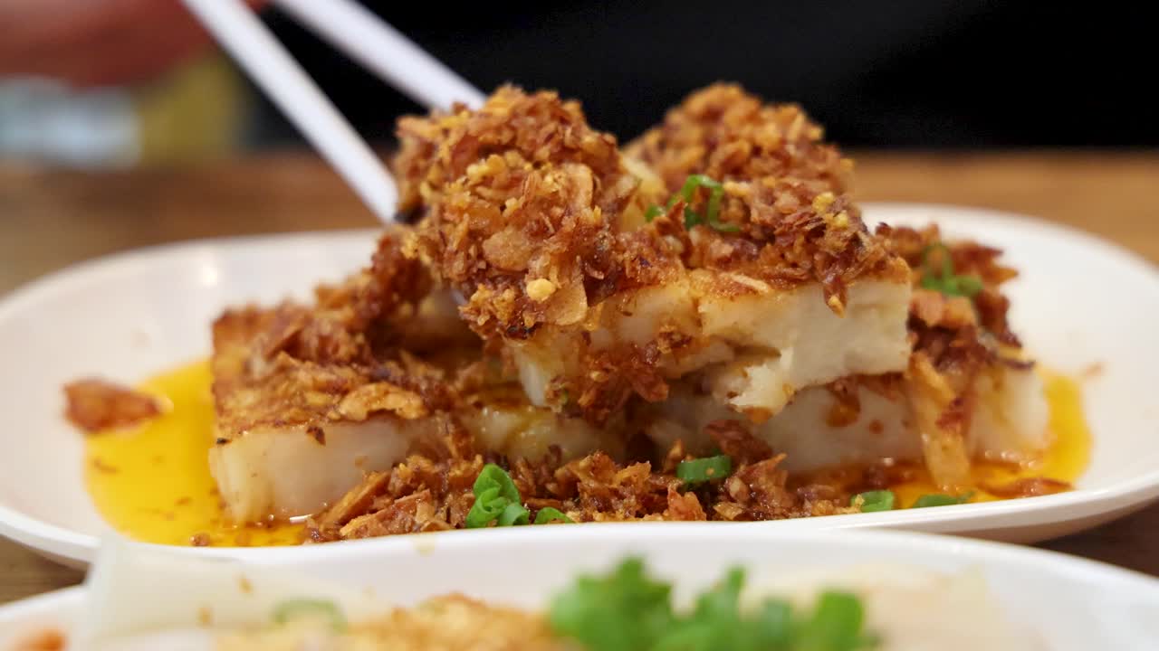 Chopsticks pick up a piece of fried turnip cake garnished with crispy fried shallots, served on a white plate in a well-lit indoor setting