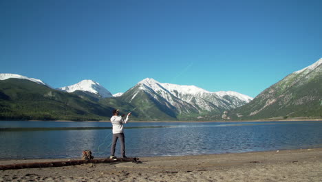 wide shot of a fly fisherman casting his lure into a deep blue lake in front of gorgeous, snow-capped mountains