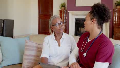 senior woman with tablet talking to nurse in living room, discussing care, at home