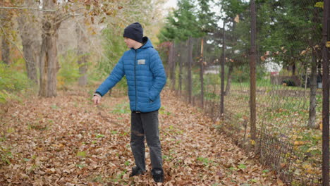 close-up of child in blue jacket and black beanie playfully scattering dry autumn leaves on the forest floor with stick, creating a lively and joyful fall outdoor scene