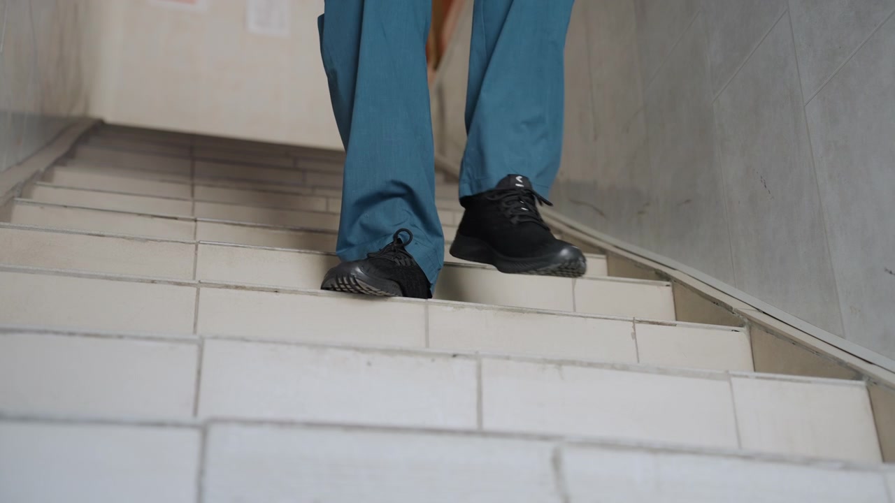 White paramedic descending tiled home stairs, empty stairwell then cautious footsteps, black shoes in blue scrubs, narrow corridor and worn treads, careful footing and safety focus, responder arrival