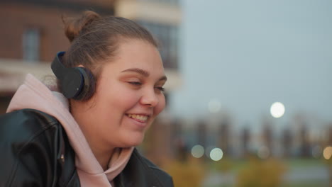 close up of cheerful girl seated outdoors putting on headphone and reaching for phone on wooden table, dressed in hoodie and leather jacket with soft bokeh lights in blurred background
