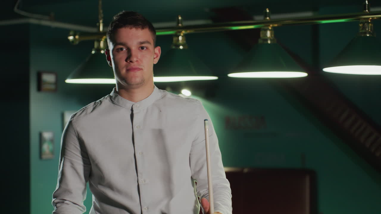 셔츠를 입은 빌리어드 선수는 희미하게 밝은 풀 홀에 서기 전에 큐 스틱을 들고 앉아 있습니다. 그는 결정적으로 다음 을 준비 할 때 상부 조명은 집중된 분위기를 니다.