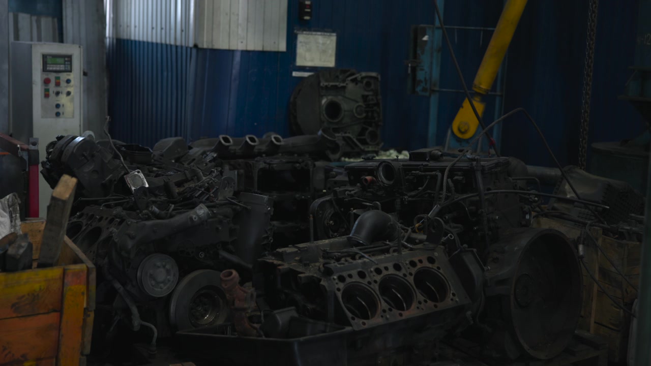 Static industrial workshop scene showing heavy disassembled engine components stacked on wooden pallets and crates, surrounded by metal equipment, grime, rust, and cool diffused light
