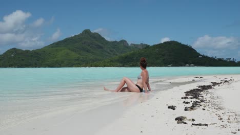 young caucasian woman in bikini and sunglasses sitting, sunbathing and relaxing on a white sand tropical beach with feet in the water and green pacific island behind in maupiti, french polynesia.