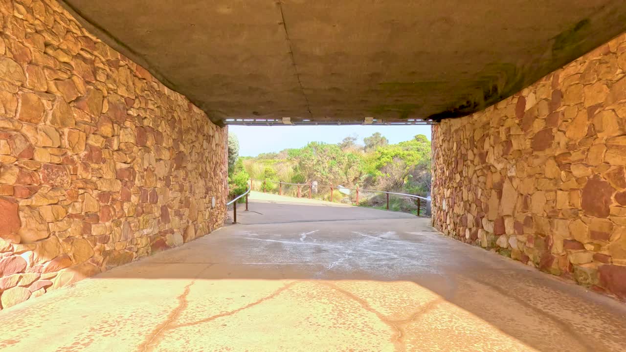 A camera moves through a sunlit stone tunnel, revealing a scenic outdoor path. The lighting creates a warm, inviting atmosphere