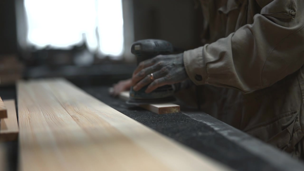 Dust-covered hands hold orbital sander flat on long pine boards over workshop bench, showing careful surface smoothing, natural light, tools, and textured wood grain detail, with sander, woodworker