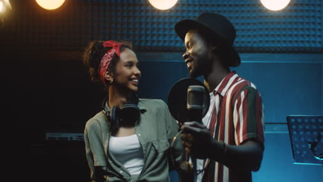 portrait of the multiethnic stylish young man and woman, singers of the duo looking at each other and then smiling cheerfully to the camera in the dark sound studio during recording