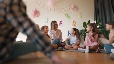 Ein-Mädchen-Mit-Bob-Frisur-Und-Brille-In-Einem-Weißen-Hemd-Kommuniziert-Mit-Ihren-Schülern,-Während-Es-In-Einem-Club-Zur-Vorbereitung-Der-Kinder-Auf-Die-Schule-Auf-Einem-Speziellen-Kissen-Auf-Dem-Boden-Sitzt