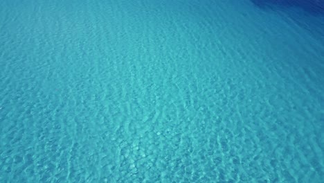 Static-drone-shot-looking-down-on-bright-blue-wavy-ocean-with-sunlit-seabed