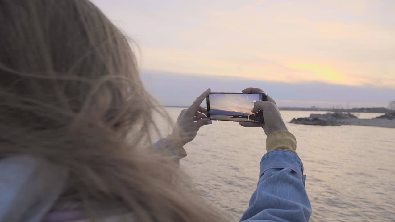 Mujer joven tomando una foto del atardecer en la costa con su teléfono.