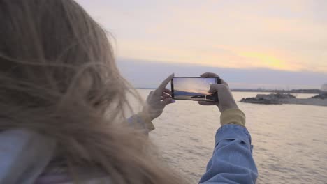 mujer joven tomando una foto de puesta de sol en la orilla del mar en el teléfono