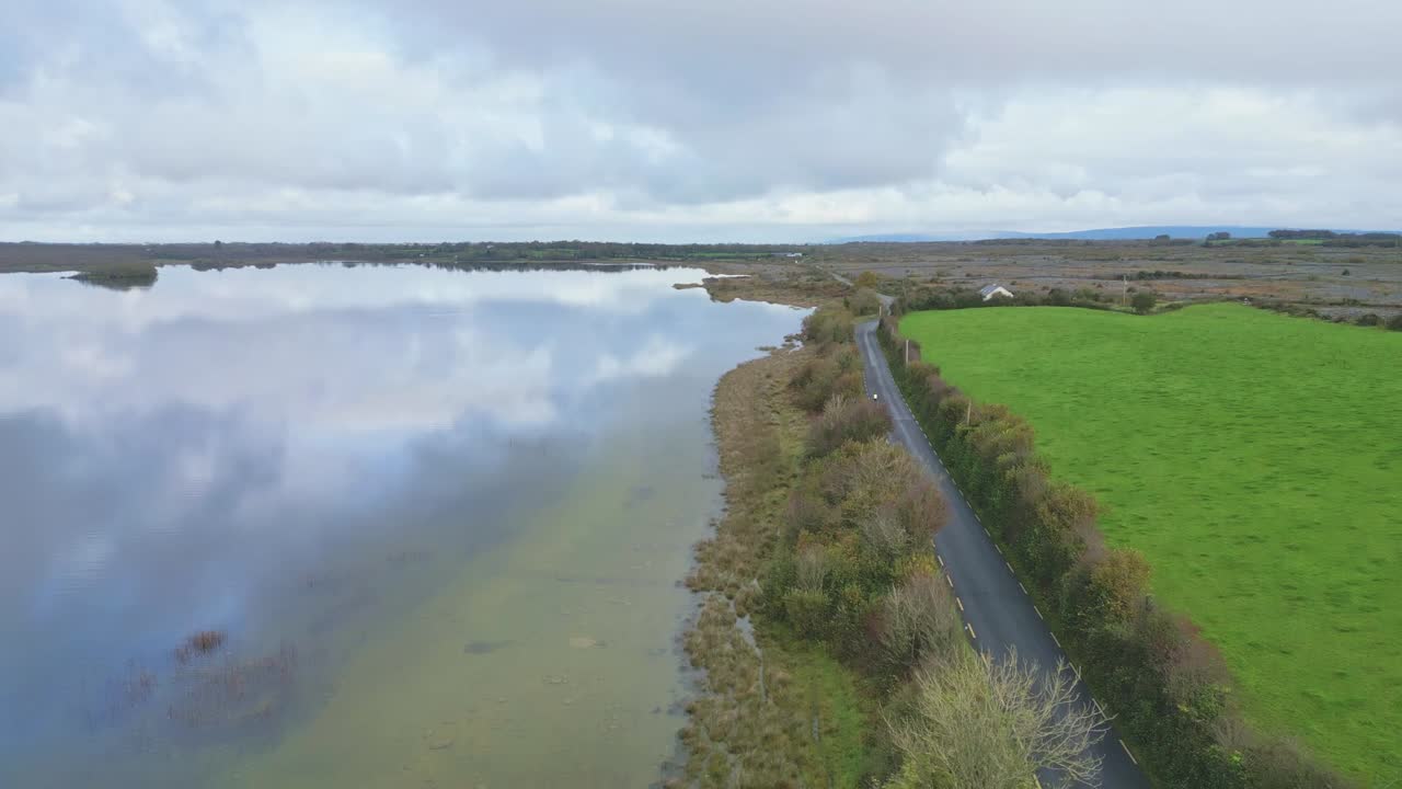 Aerial view of the Burren in Ireland, showing a winding road along the water’s edge, lush green fields, and serene rural scenery under a cloudy sky