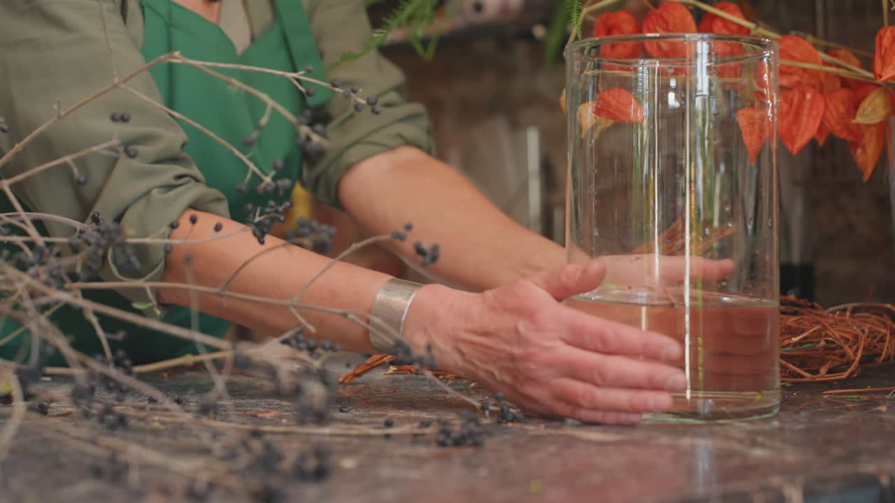 Květinářka plnící průhlednou skleněnou vázu vodou, ruce pevně na pultu, roztroušené zbytky větviček, nástroje na stříhání poblíž, pečlivá příprava podzimního aranžmá, odraz na skle a detail jemného vlnění.