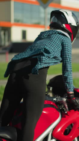 a female motorcyclist, wearing a checkered shirt and canvas shoes, rides a power bike standing, a blue truck is visible ahead of her with parked cars in the background on an open road