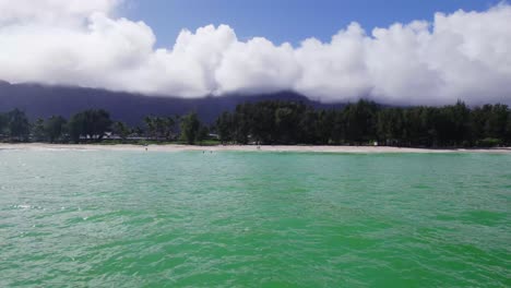 drone-footage-panning-across-a-white-sandy-beach-with-turquoise-water-in-the-foreground-and-lush-greenery,-purple-mountains-and-blue-sky-with-white-clouds-in-the-background-Oahu-Hawaii