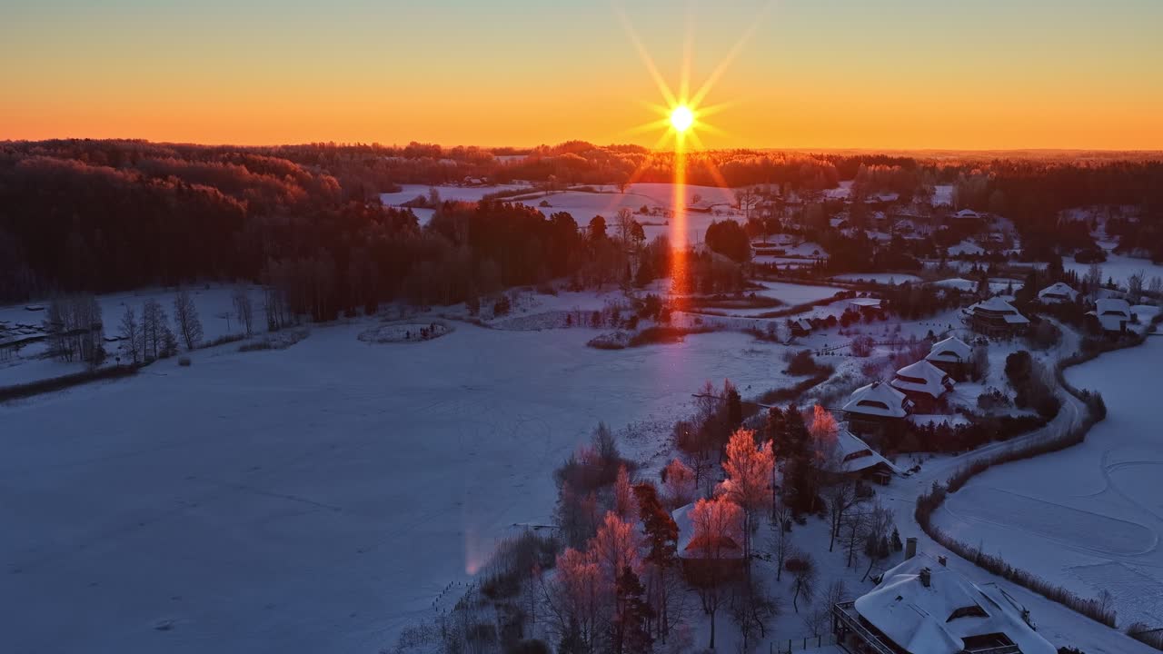 Drooninäkymä talviauringonlaskusta jäätyneen järven ja lumisten peltojen yllä lähellä maalaiskylää, kultaisen tunnin auringonsäteiden ja värikkään taivaan kera hiljaisen maaseudun yllä
