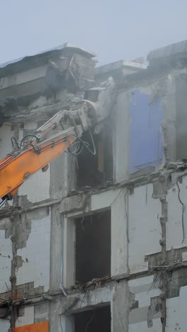 excavator long claw working in a dusty cloud. demolishing machinery destroys the roof of an old house. debris falling heavily from the roof. vertical video