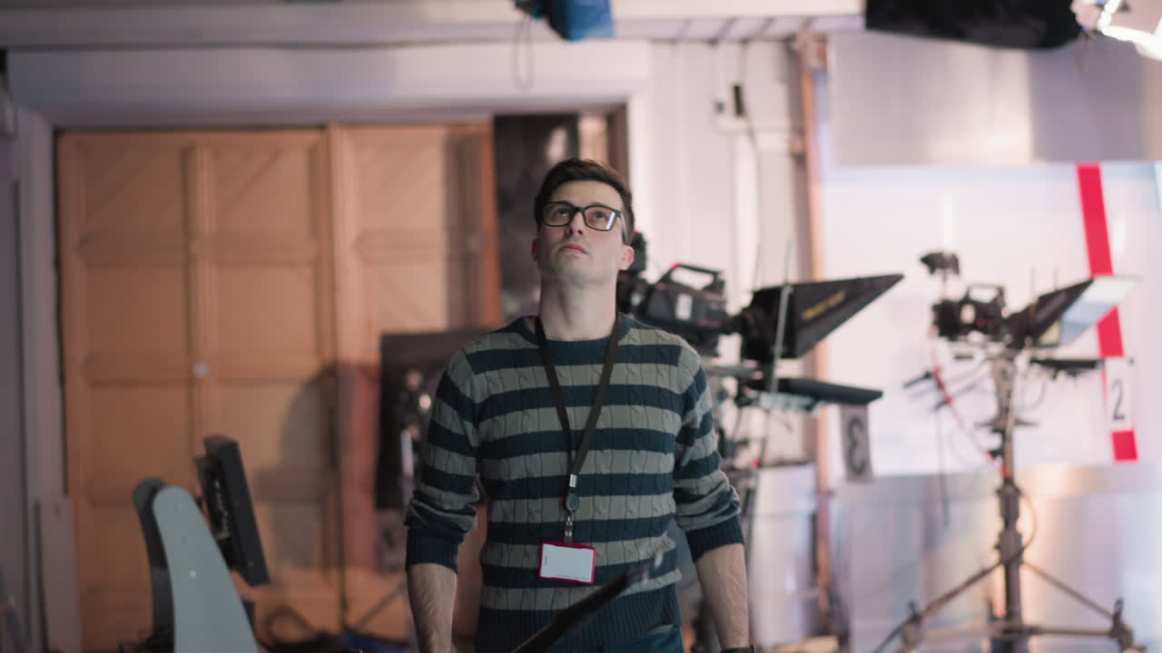 Technician in broadcast studio with boom pole adjusting lighting. Focused on controlling media equipment in professional setup with visible camera and lighting in background