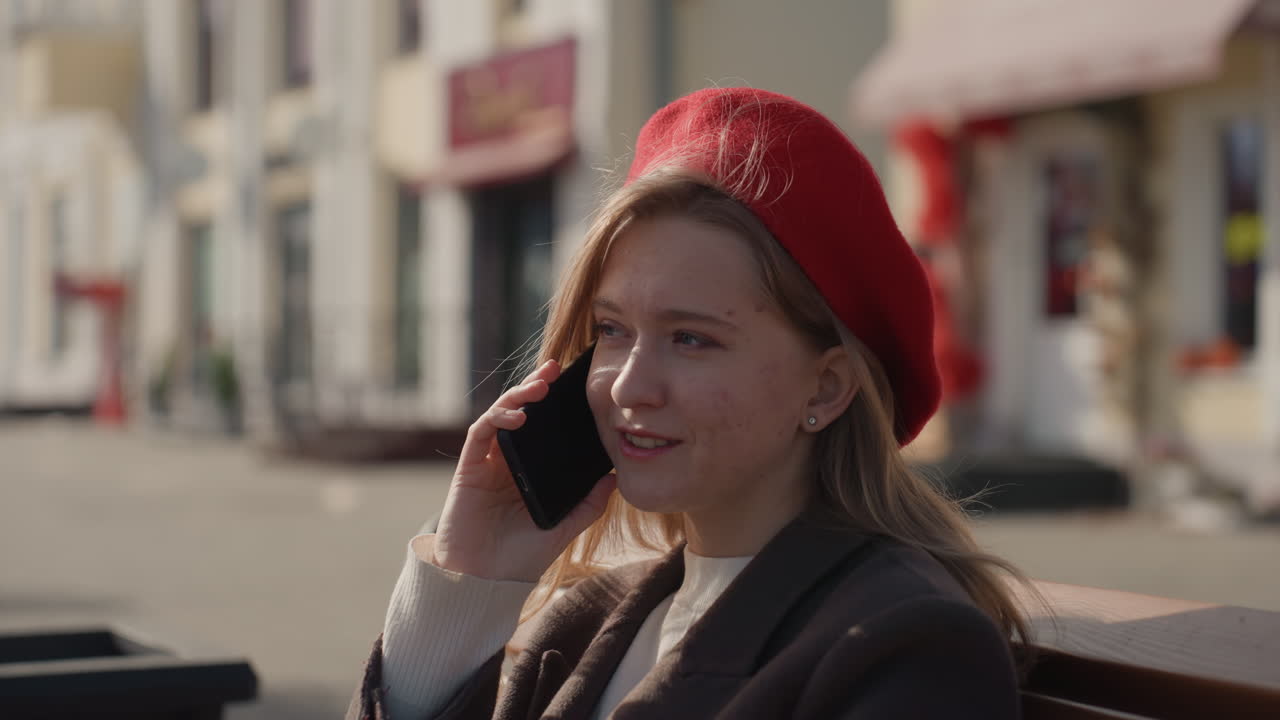 Jovem mulher caucasiana em um banco, em uma rua urbana ensolarada, com boina vermelha, falando ao smartphone, sorrindo e reflexiva, com folhagem de outono e vitrines ao fundo, transmitindo uma vibe de escritora relaxada, um momento de estilo de vida espontâneo.