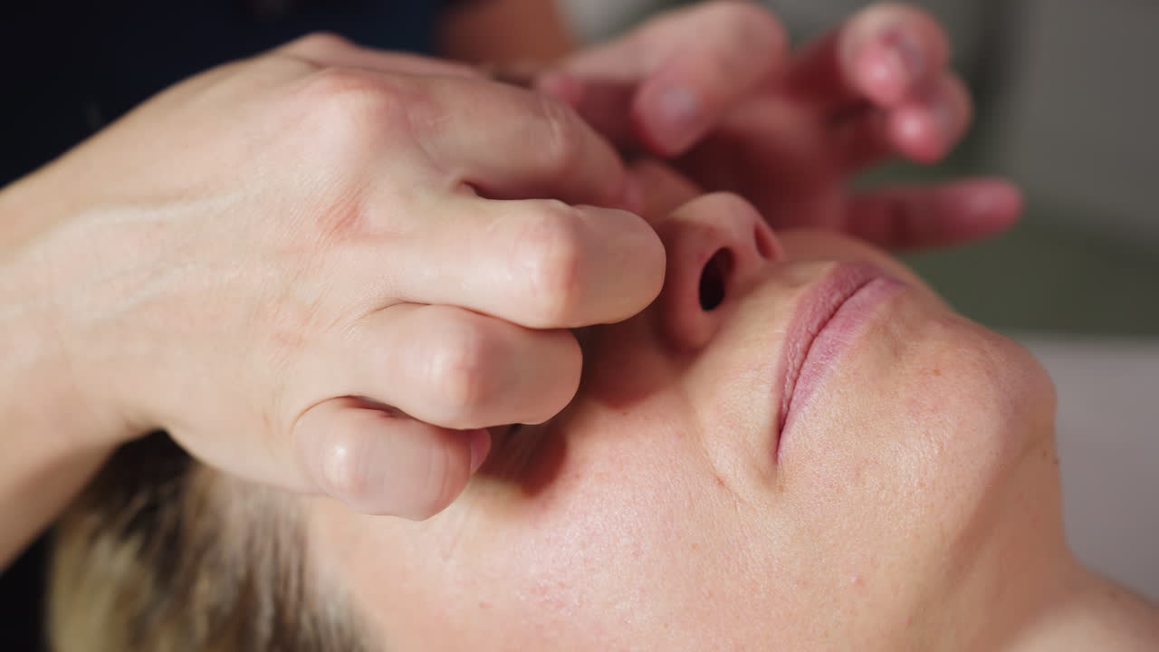 Close up of participant lying face up on massage bed as bodywork therapist uses gentle circular motions to massage brows and forehead, smoothing skin, releasing tension in spa environment