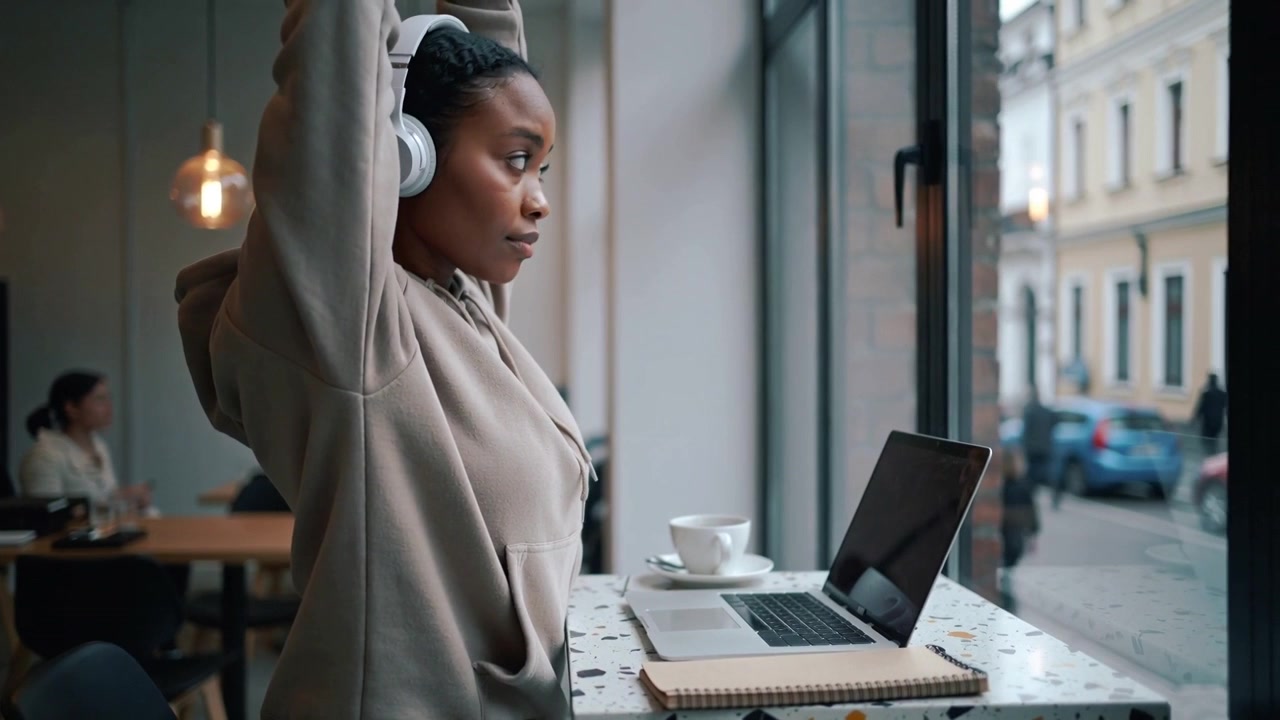 Putting on white headset woman in hoodie at cafe, stretching during open laptop break
