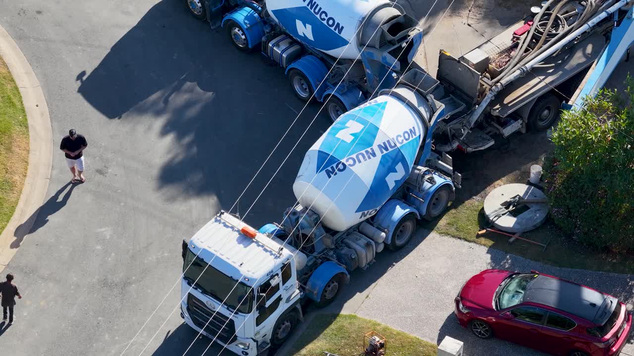Concrete mixer truck operates on suburban street, person walks nearby, bright daylight, aerial perspective
