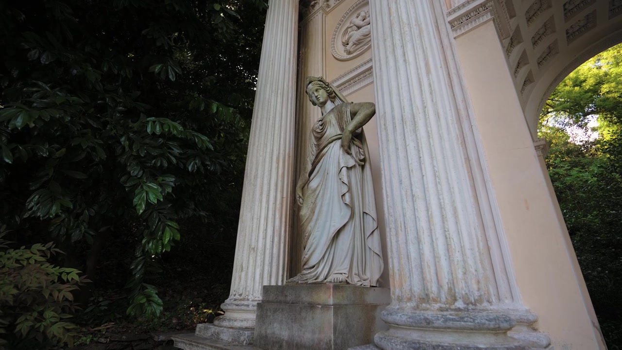Ripresa dal basso di una statua allegorica classica in marmo che si erge tra grandi colonne scanalate incorporate nella facciata di una storica struttura ad arco trionfale.