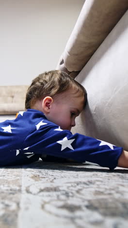 toddler boy lies on the floor reaching his hand under the sofa. baby wants to get something from under the couch. vertical video