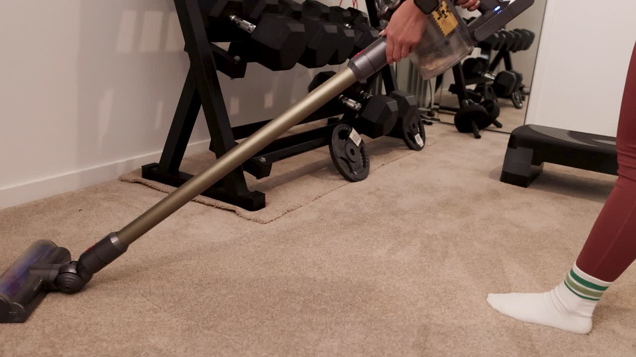 A person vacuums a carpeted home gym with weights, using a cordless vacuum cleaner