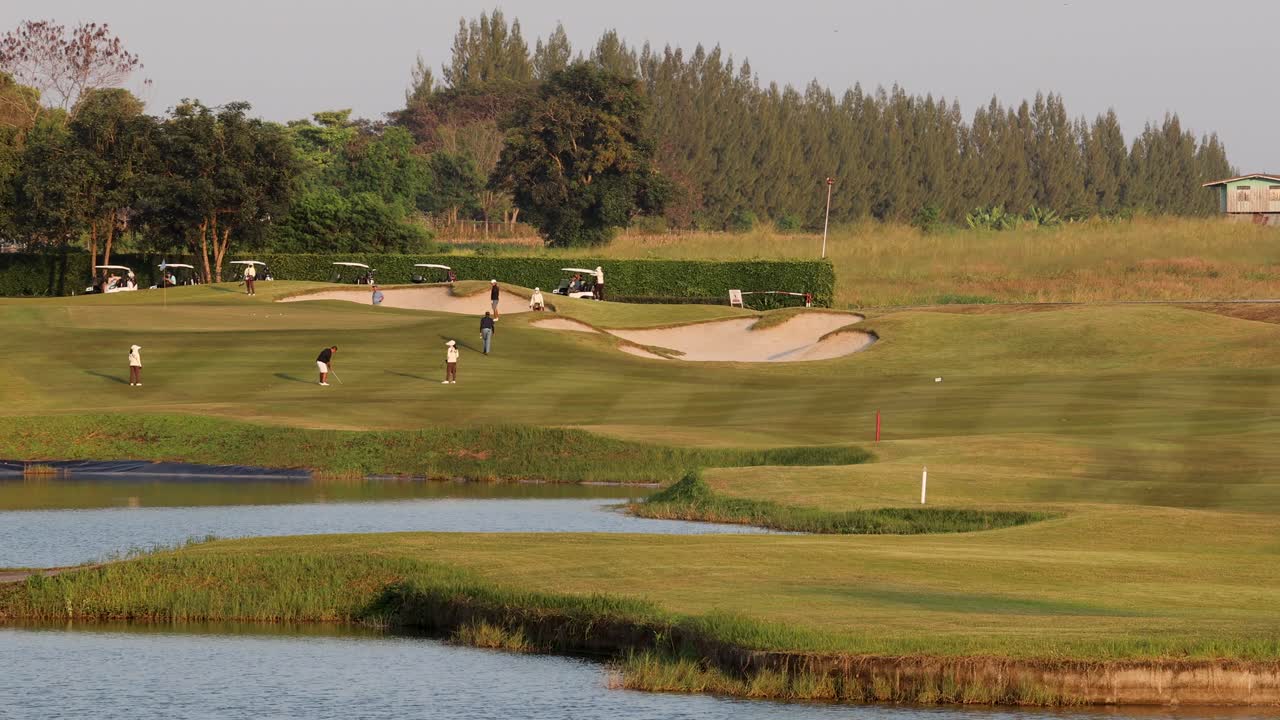 Golfers walk and play on lush course with lake, mountains, warm sunset lighting, wide shot