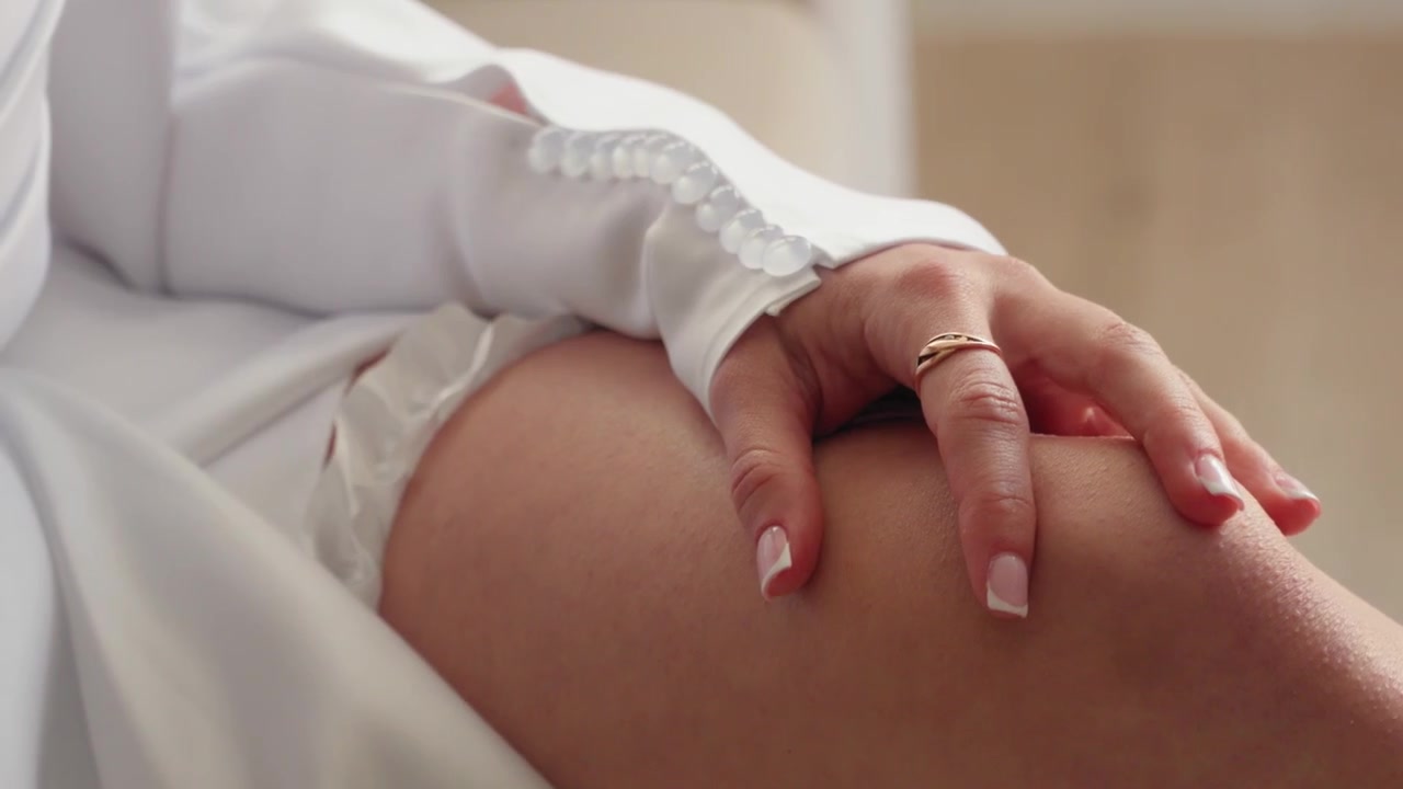 Primo piano della mano curata di una sposa che sistema una delicata giarrettiera bianca sotto la manica del suo abito da sposa, catturato con luce brillante e naturale, con dettagli morbidi ed eleganti e un focus femminile.