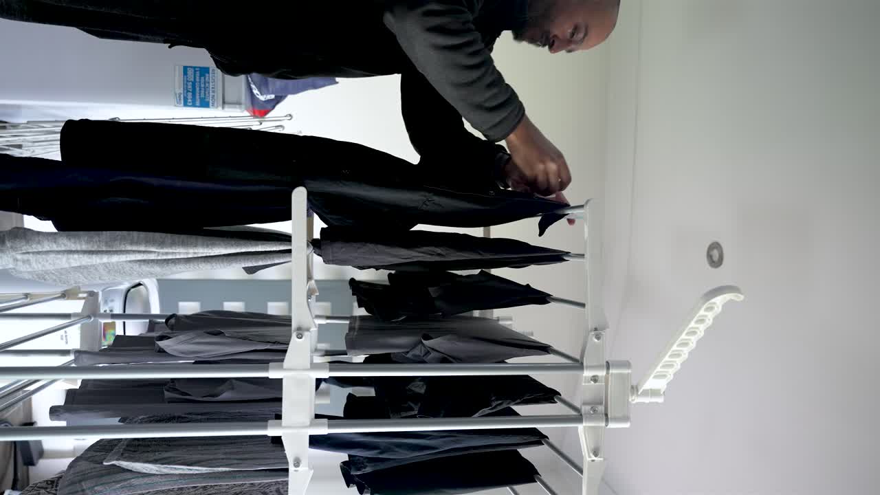 Uomo asiatico-britannico che stende i vestiti su uno stendibiancheria da interno, incarnando il concetto di bucato al chiuso e faccende domestiche quotidiane