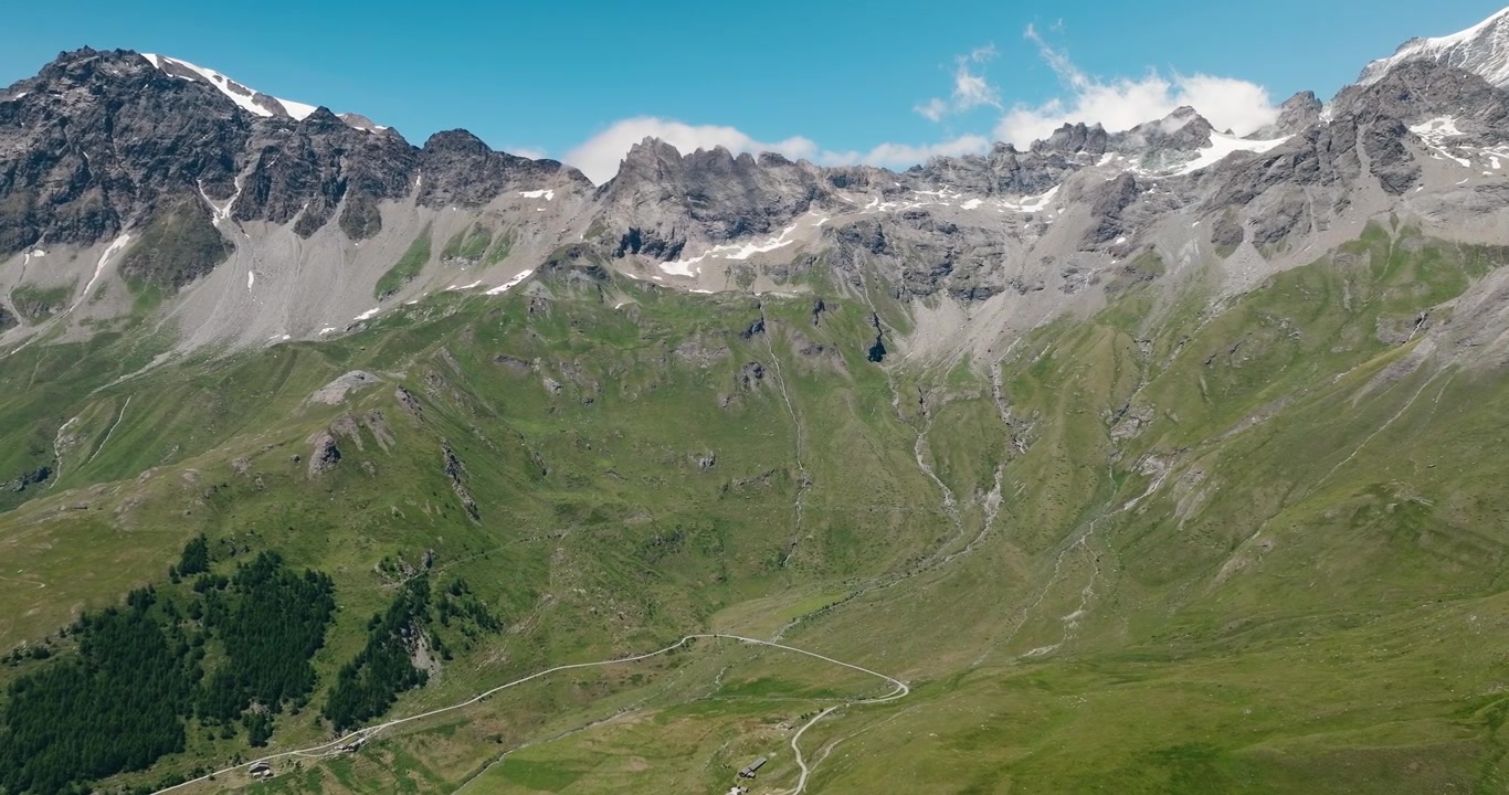 Vista panoramica da una lussureggiante valle alpina che guarda verso imponenti montagne di granito con cime innevate sotto un cielo estivo azzurro e cristallino, mostrando una bellezza naturale mozzafiato