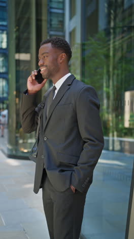 vertical video shot of smiling young businessman wearing suit talking on mobile phone standing outside offices in the financial district of the city of london uk shot in real time 3