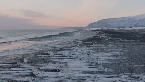 Aerial-landscape-view-of-ocean-waves-crashing-on-to-diamond-beach,-covered-in-snow,-at-dusk